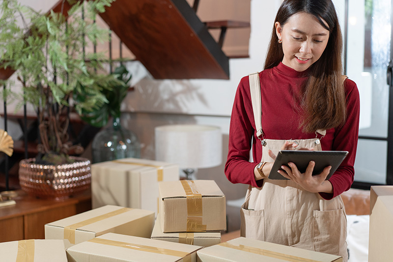 A woman is surrounded by boxes. She is looking at an iPad, checking inventory in her store.