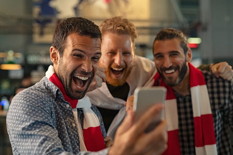 Three men are looking at a smartphone, watching a football match and cheering.